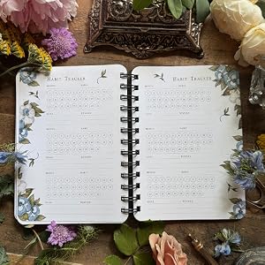 An open spiral-bound planner or journal spread on a rustic wooden surface, surrounded by flowers and a decorative tray. The pages are white with floral illustrations along the left margin and printed tables under the heading 'HARD TRAFFICKS'.
