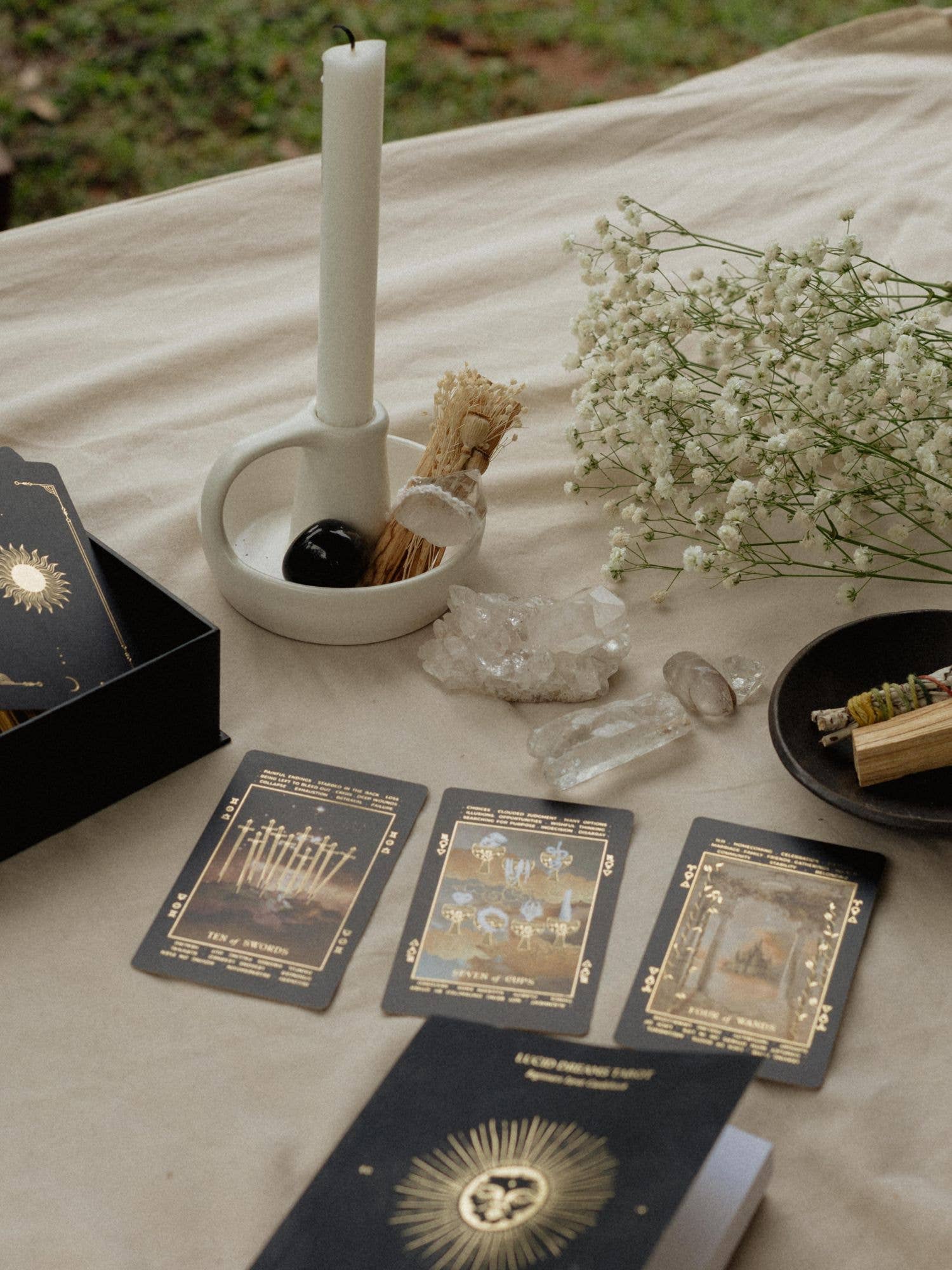A styled tarot card reading setup on a beige fabric surface outdoors, featuring several black and gold oracle or tarot cards, a white candle in a white holder, crystals, dried grass, and baby's breath flowers.