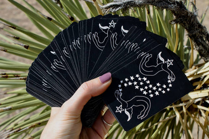 Black tarot cards with white symbols held by a hand against a natural background DIVINA Tarot Deck by Mary Elizabeth Evans featuring elegant monochrome pen and ink artwork with celestial symbolism and a 78-card indie tarot deck.