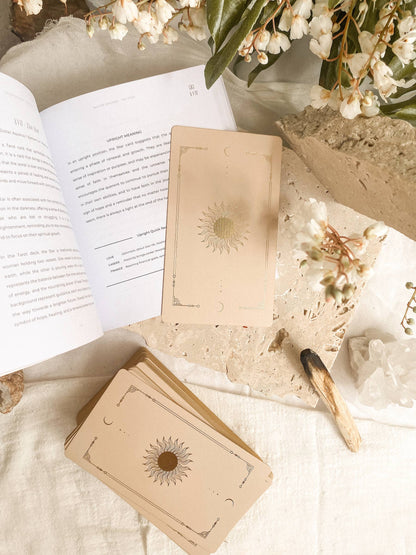 A styled flat-lay of a tarot card deck with beige cards featuring sunburst and moon designs, an open book, dried flowers, crystals, and a match on a soft cream fabric background. Lucid Dreams Beginner's Tarot Beige Edition IV by ST. SOLEIL with gold foil cards, matte gold edges and guidebook