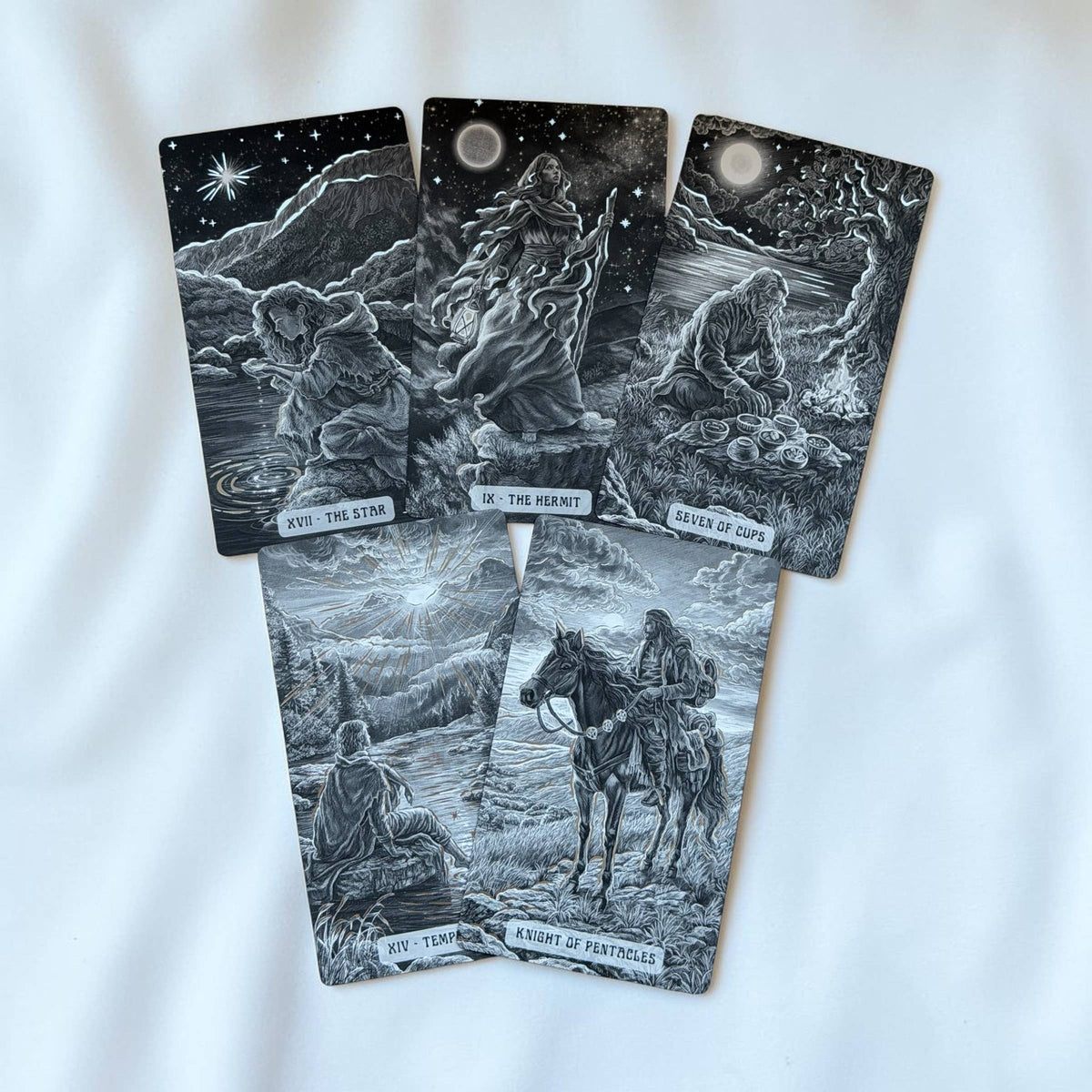 Five tarot cards with black and white gothic illustrations are arranged on a white fabric background. The cards feature detailed fantasy and landscape scenes with moonlit skies, mountains, figures, and a horse rider. Caminante Tarot deck with hardcover guidebook featuring silver foil tarot cards and matte black edges

