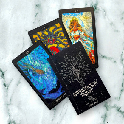 Four tarot cards and the Aspdron Tarot deck box arranged on a white and green marble surface. The box is black with a silver tree design and the title 'ASPDON TAROT'. Asphodelon Tarot by Asphodelon Studio featuring hand-painted Greek mythology artwork, gilded edges, premium matte cards and a deluxe gold foil presentation box.