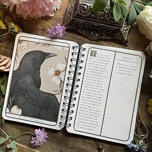 An open spiral-bound journal laid on a rustic wooden surface, surrounded by pink and white flowers. The left page shows a black raven illustration with floral details and the title 'Intelligence.