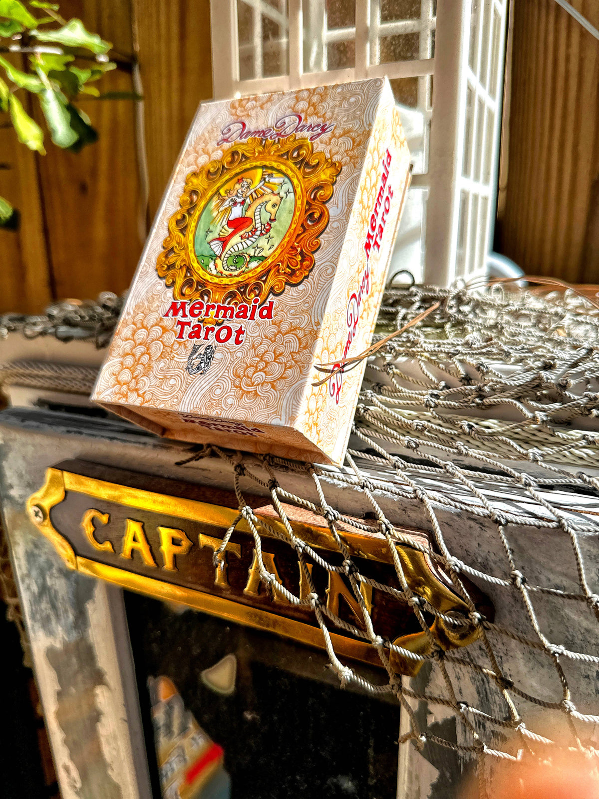 A boxed deck of Mermaid Tarot cards by Dreamy Dreams sits on a rustic nautical-themed surface with a Captain's plaque and netting in the background.