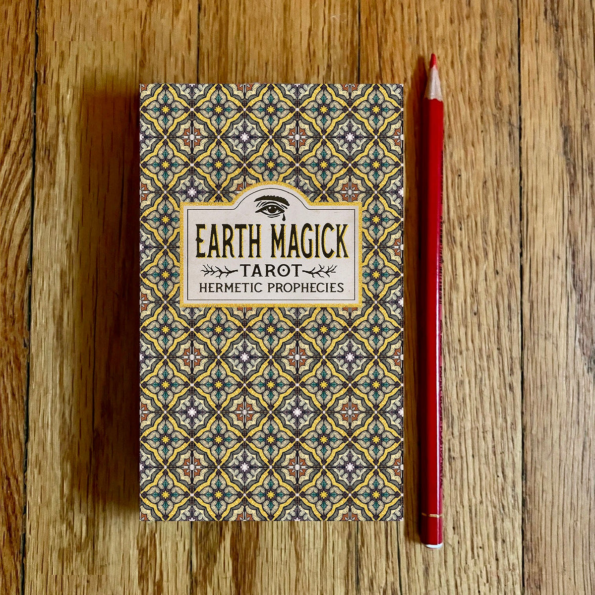 A tarot deck box labeled 'Earth Magick Tarot Hermetic Prophecies' sits on a wooden surface next to a red pencil. The box features an intricate geometric pattern in blue, yellow, and gray.