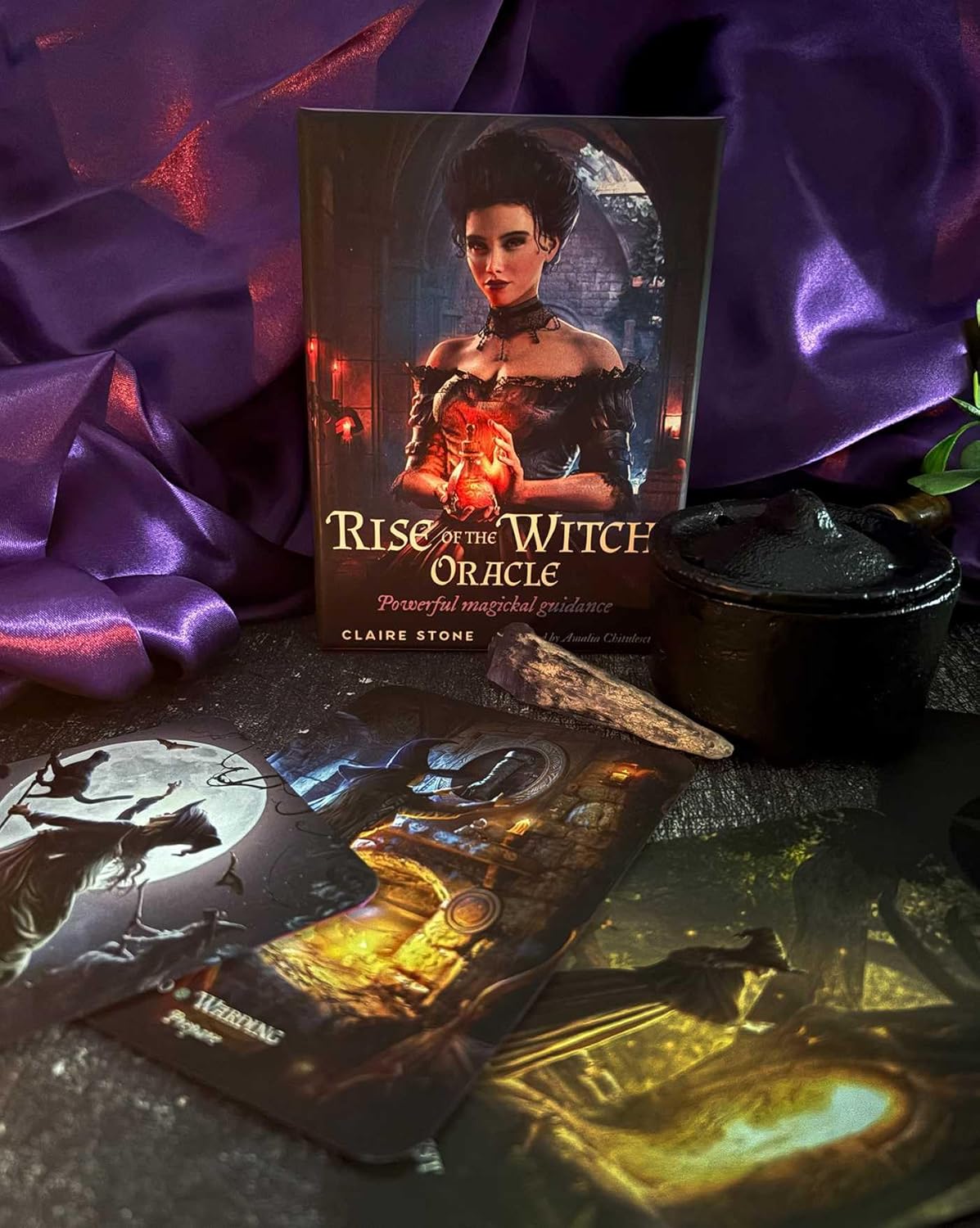 The book 'Rise of the Witch Oracle' by Claire Stone is displayed upright, surrounded by its illustrated oracle cards, a black cauldron, and draped purple silk fabric.