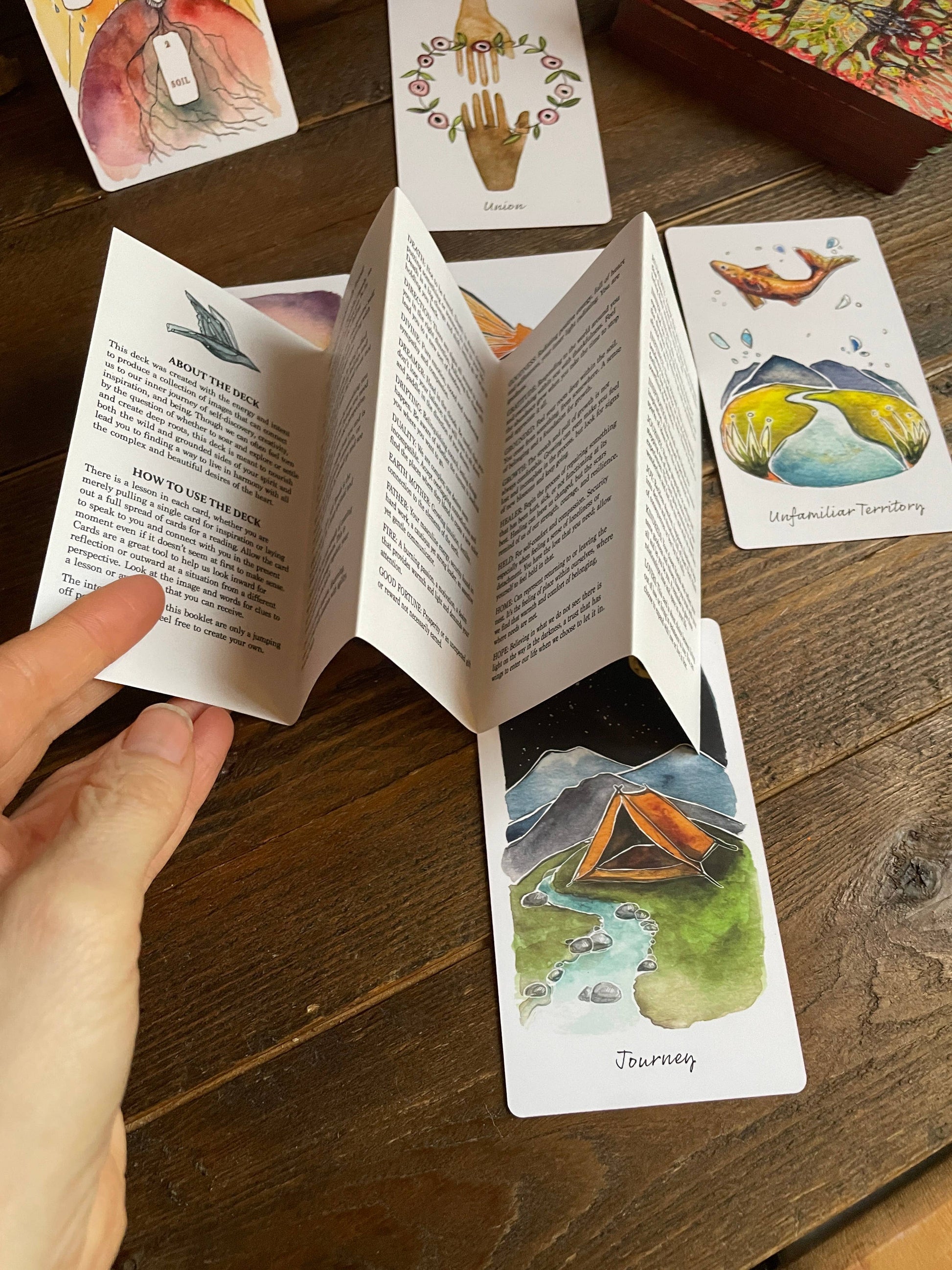 Card with a hand-drawn illustration of a tent and nature, held open by a hand on a wooden surface with other cards in the background.