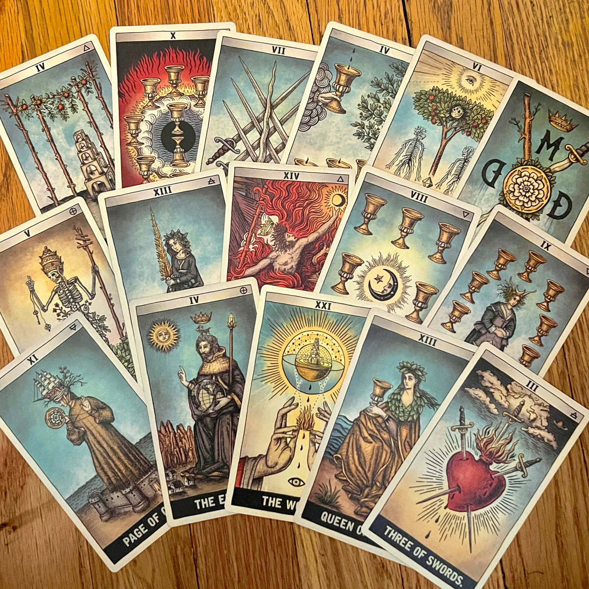 A fan arrangement of multiple Rider-Waite tarot cards on a wooden tabletop, showing illustrated faces and symbols including Page, Knight, Queen, King, The World, Three of Swords, and The Hanged Man.