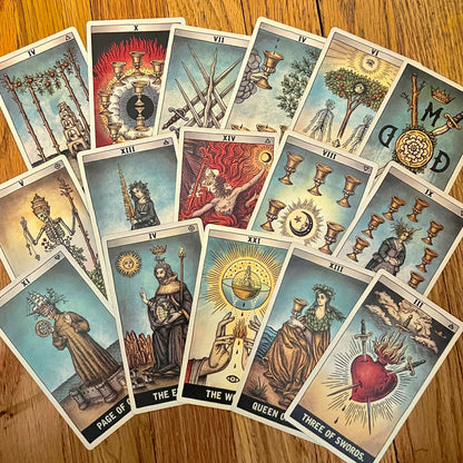 A fan arrangement of multiple Rider-Waite tarot cards on a wooden tabletop, showing illustrated faces and symbols including Page, Knight, Queen, King, The World, Three of Swords, and The Hanged Man.