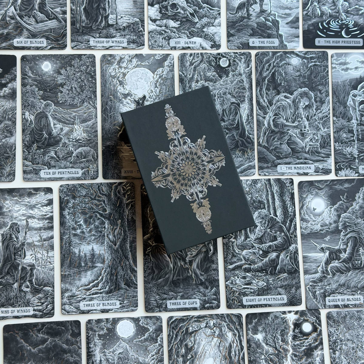 A tarot deck box with a dark charcoal cover and ornate silver filigree sits on top of a spread of illustrated tarot cards showing the 'Three of Pentacles', 'Three of Cups', and 'Three of Swords' illustrations. Caminante Tarot deck with hardcover guidebook featuring silver foil tarot cards and matte black edges

