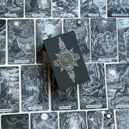 A tarot deck box with a dark charcoal cover and ornate silver filigree sits on top of a spread of illustrated tarot cards showing the 'Three of Pentacles', 'Three of Cups', and 'Three of Swords' illustrations. Caminante Tarot deck with hardcover guidebook featuring silver foil tarot cards and matte black edges

