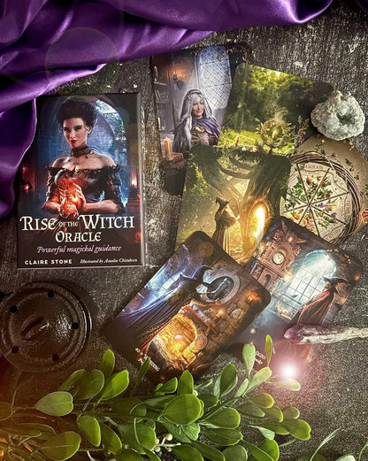 A styled flat lay of the Rise of the Witch Oracle card deck by Claire Stone, showing several illustrated oracle cards with fantasy and witch-themed artwork, a purple fabric drape, green leaves, a metal weight, and a small stone on a dark textured surface.