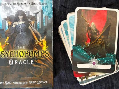 The Pychopomps Oracle card deck shown with its illustrated box on the left and several cards fanned out on the right, placed on a dark fabric surface. The artwork features a dark gothic figure in a boat with red, teal, and black tones.