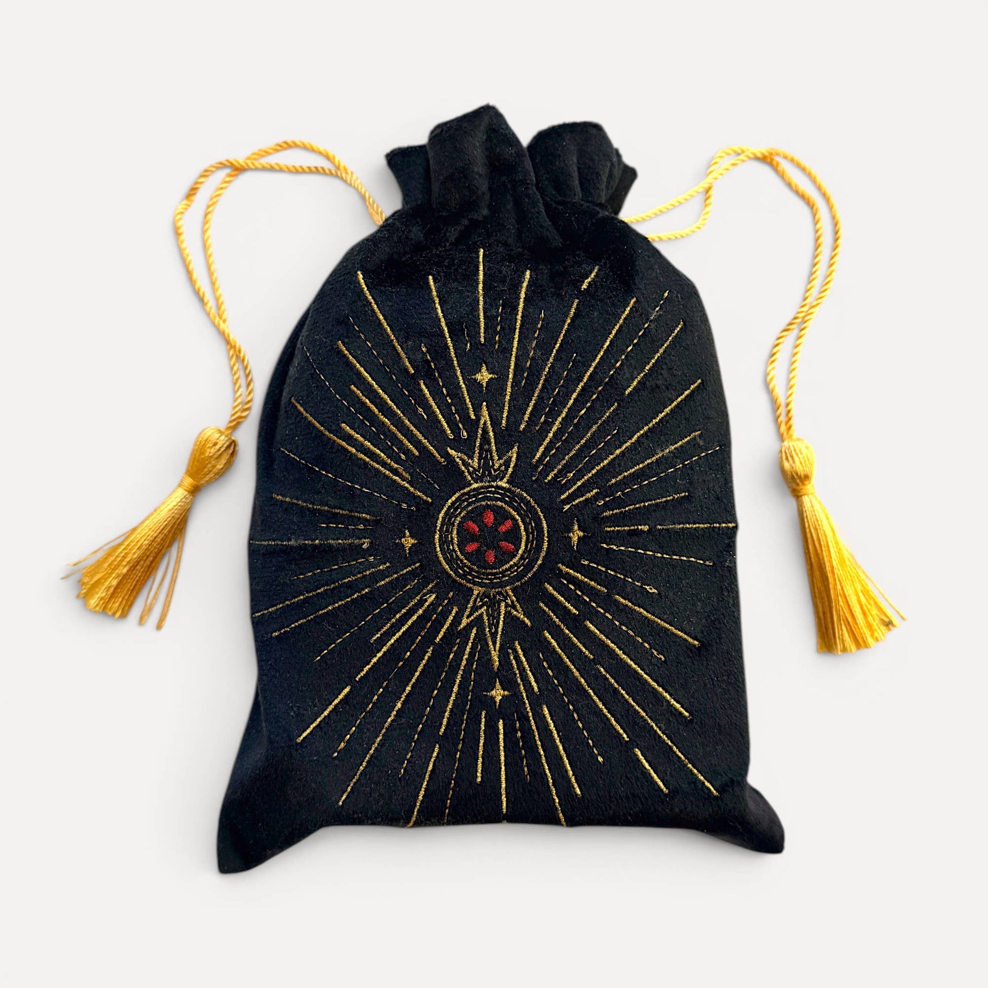 A black velvet drawstring pouch with gold celestial sunburst embroidery and a red central emblem, featuring gold tassel ties, set against a light gray background.