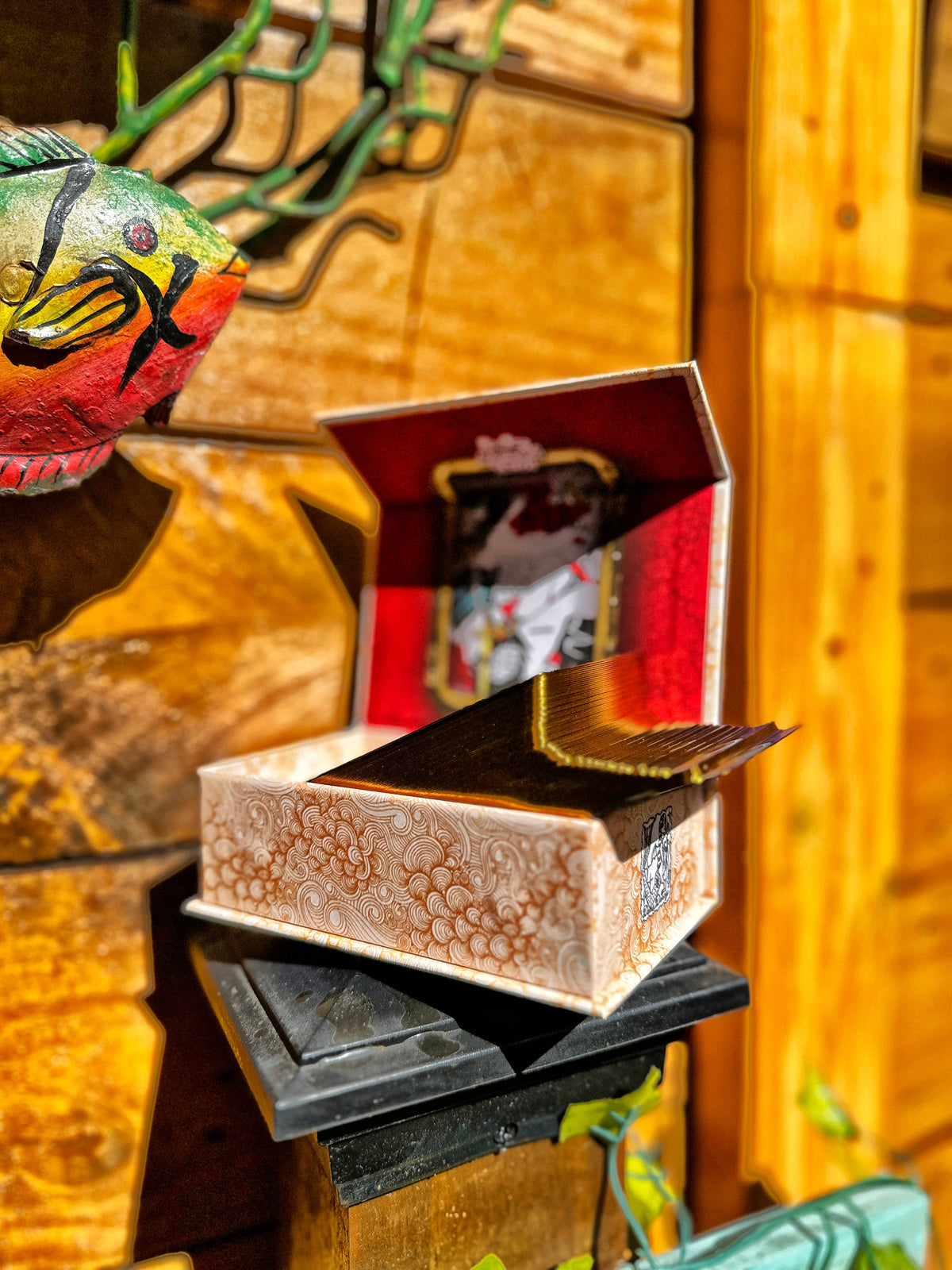 A decorative patterned box with a red interior lid, shown open on a black pedestal against a warm wooden background. A colorful fish-shaped decorative object is partially visible on the left. Gold-gilded card edges on the Dame Darcy Mermaid Tarot Ultra Gold Edition