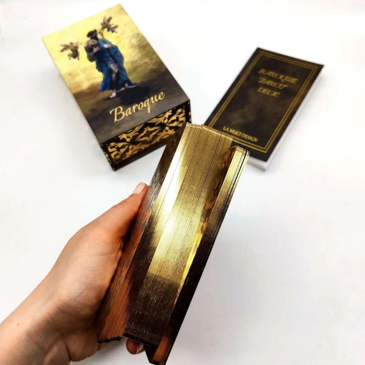 Hand holding a decorative book with a gold cover, next to its packaging on a white background Baroque tarot La Muci