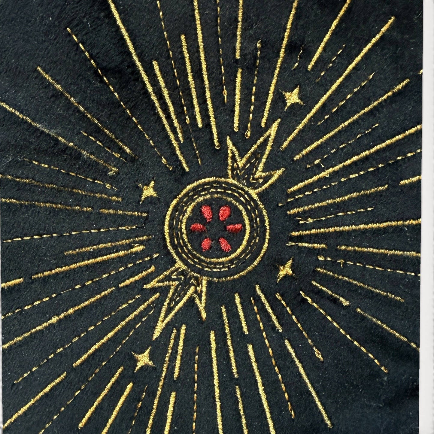 A close-up of a black fabric patch featuring a gold embroidered sunburst design with a central red and gold circular motif.