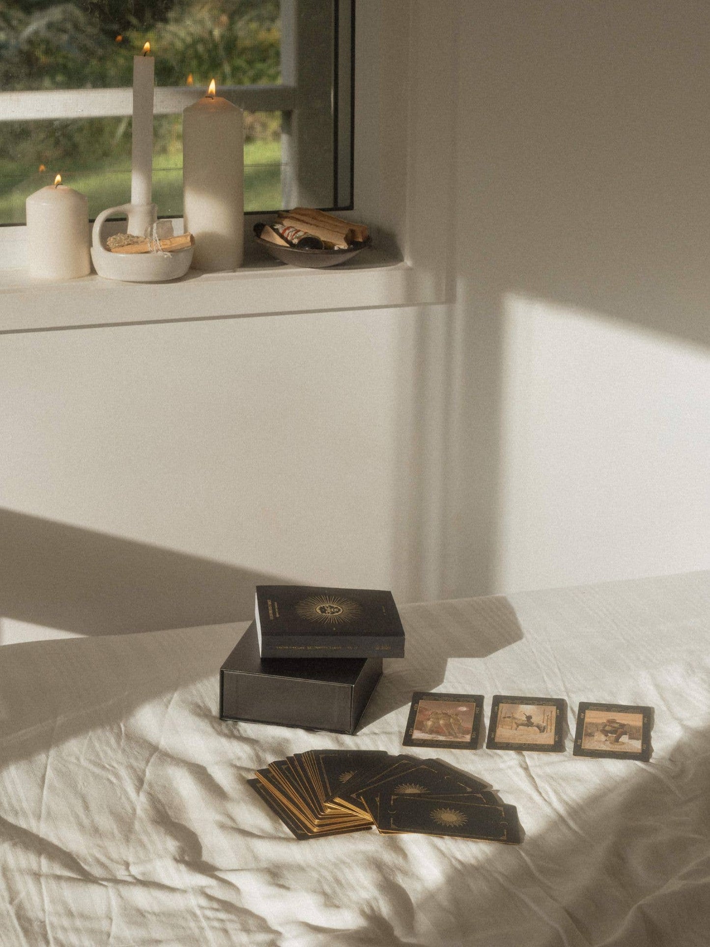 A tarot card deck set arranged on a white bed in a softly lit room, with the box open, cards fanned out, three cards laid face up, and candles on a windowsill in the background. Lucid Dreams Beginner's Tarot Black Eclipse Edition IV by ST. SOLEIL featuring matte black cards with gold foil details, premium linen finish and magnetic keepsake box.