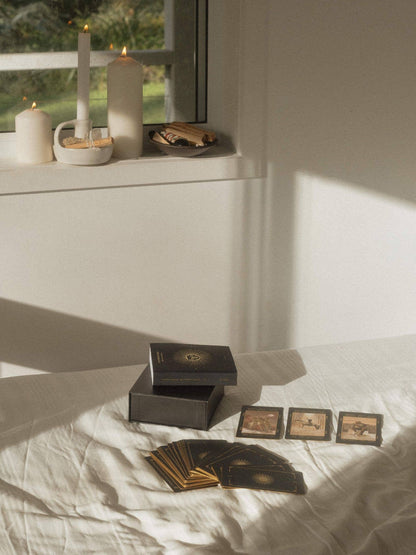 A tarot card deck set arranged on a white bed in a softly lit room, with the box open, cards fanned out, three cards laid face up, and candles on a windowsill in the background. Lucid Dreams Beginner's Tarot Black Eclipse Edition IV by ST. SOLEIL featuring matte black cards with gold foil details, premium linen finish and magnetic keepsake box.
