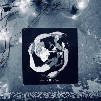 Decorative coaster with a black and white illustration of mermaids on a textured surface with fairy lights. Dark Days Tarot by Wren McMurdo Brignac featuring square black-and-white tarot cards inspired by the lunar cycle with a 180-page guidebook and collector's box.