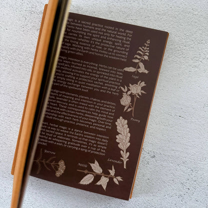 Brown book with engraved plant illustrations and text on a textured white surface Grimoire Bouchette and Co Oneness Emporium