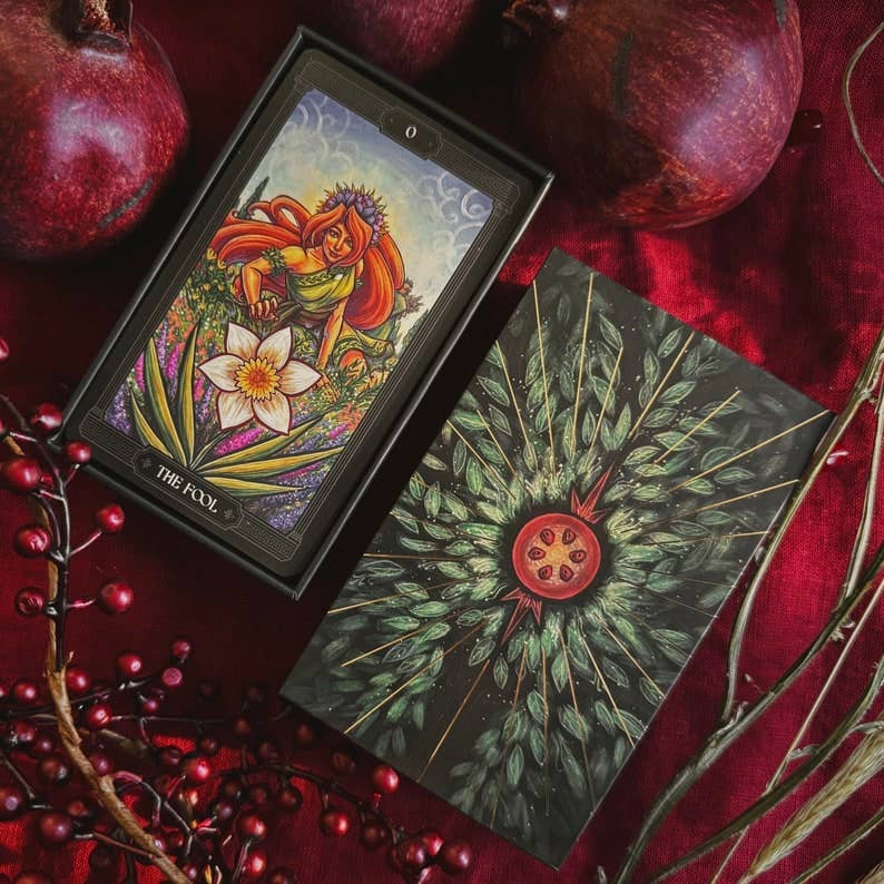 A tarot card deck box and card laid on a rich red fabric, surrounded by whole pomegranates and red berries. The box shows the illustrated front with a fairy and floral artwork, and the card beneath displays a dark back design with radiating green leaves and a red pomegranate motif. Asphodelon Tarot by Asphodelon Studio featuring hand-painted Greek mythology artwork, gilded edges, premium matte cards and a deluxe gold foil presentation box.