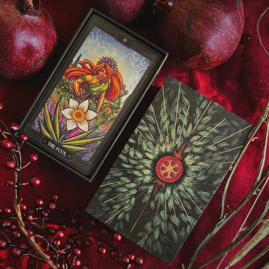 A tarot card deck box and card laid on a rich red fabric, surrounded by whole pomegranates and red berries. The box shows the illustrated front with a fairy and floral artwork, and the card beneath displays a dark back design with radiating green leaves and a red pomegranate motif. Asphodelon Tarot by Asphodelon Studio featuring hand-painted Greek mythology artwork, gilded edges, premium matte cards and a deluxe gold foil presentation box.