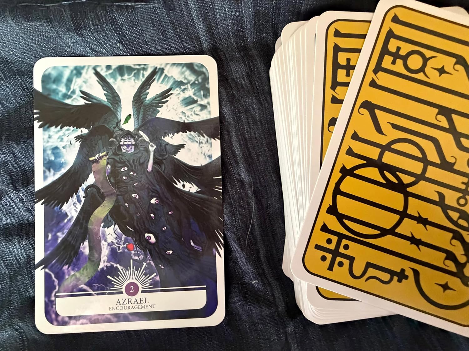 A tarot card deck laid out on a dark textured fabric surface, with one front card showing a black raven illustration and a stack of yellow-backed cards on the right.