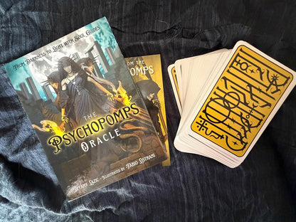 The Psychopomp's Oracle deck and box are displayed on a dark textured fabric background. The box features fantasy artwork of a woman with a scythe, and the deck of cards shows yellow-backed cards with black symbols.