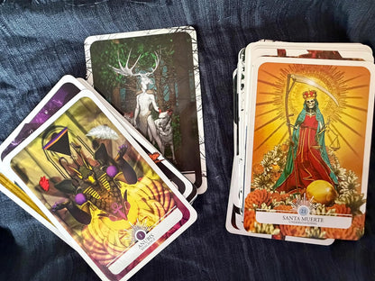 A deck of illustrated oracle or tarot-style cards arranged on dark blue fabric, with several cards fanned out and a stack showing a Santa Muerte card with a red robe, green cloak, and golden sunburst background.