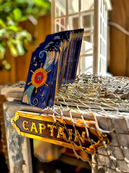 A deck of blue captain-themed playing cards with a nautical compass design, standing on a ship's wheel and net display next to a wooden 'CAPTAIN' sign.