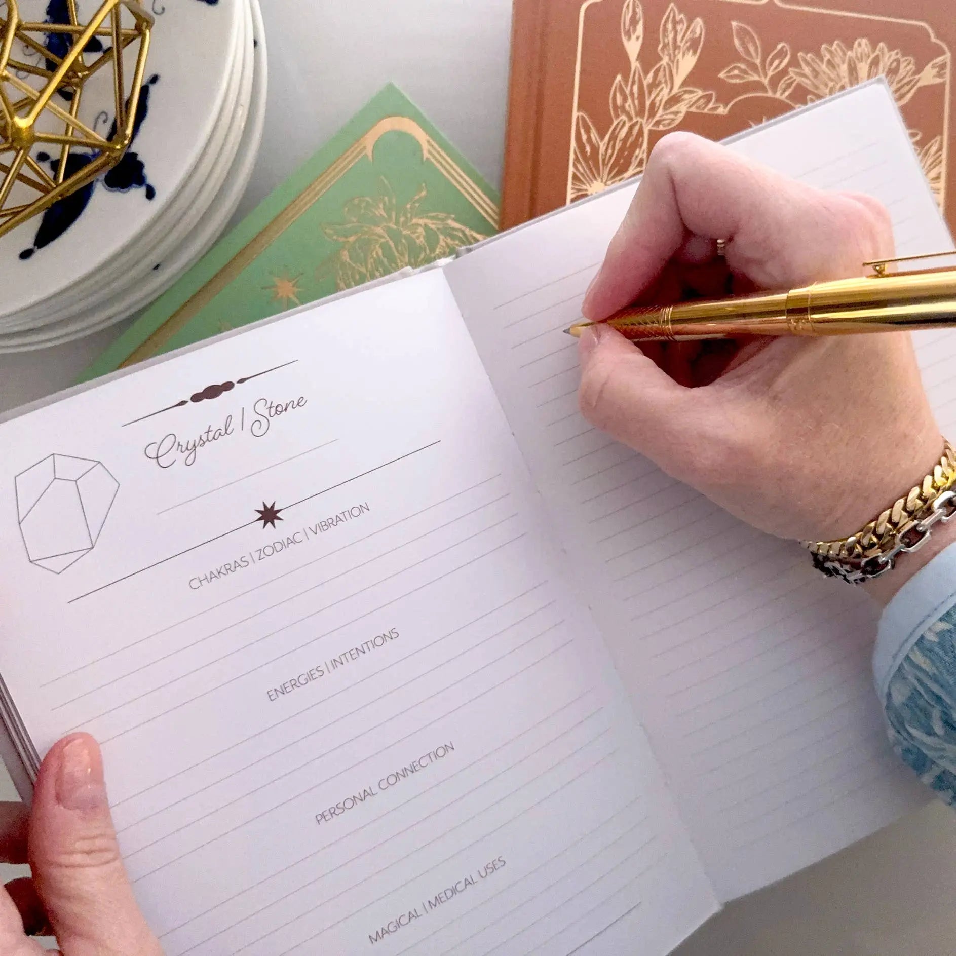 Person writing in a notebook with a gold pen on a desk with decorative items. Crystal companion journal by Bouchette and Co available from Oneness Emporium