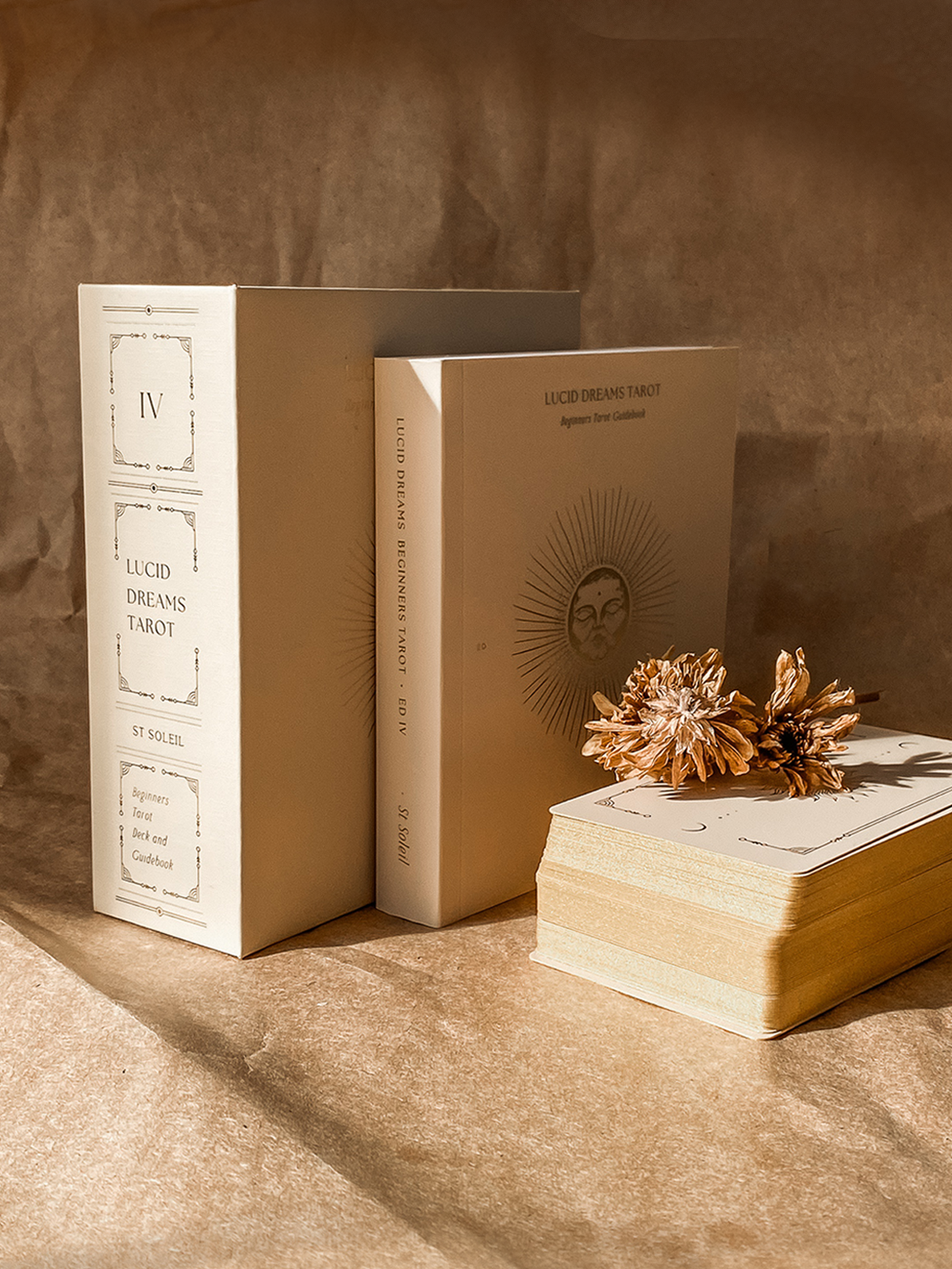 A set of cream-colored tarot card books and a matching deck box from Lucid Dreams Tarot by St. Neil, styled on a textured brown paper background with dried flowers. Lucid Dreams Beginner's Tarot Beige Edition IV by ST. SOLEIL with gold foil cards, matte gold edges and guidebook