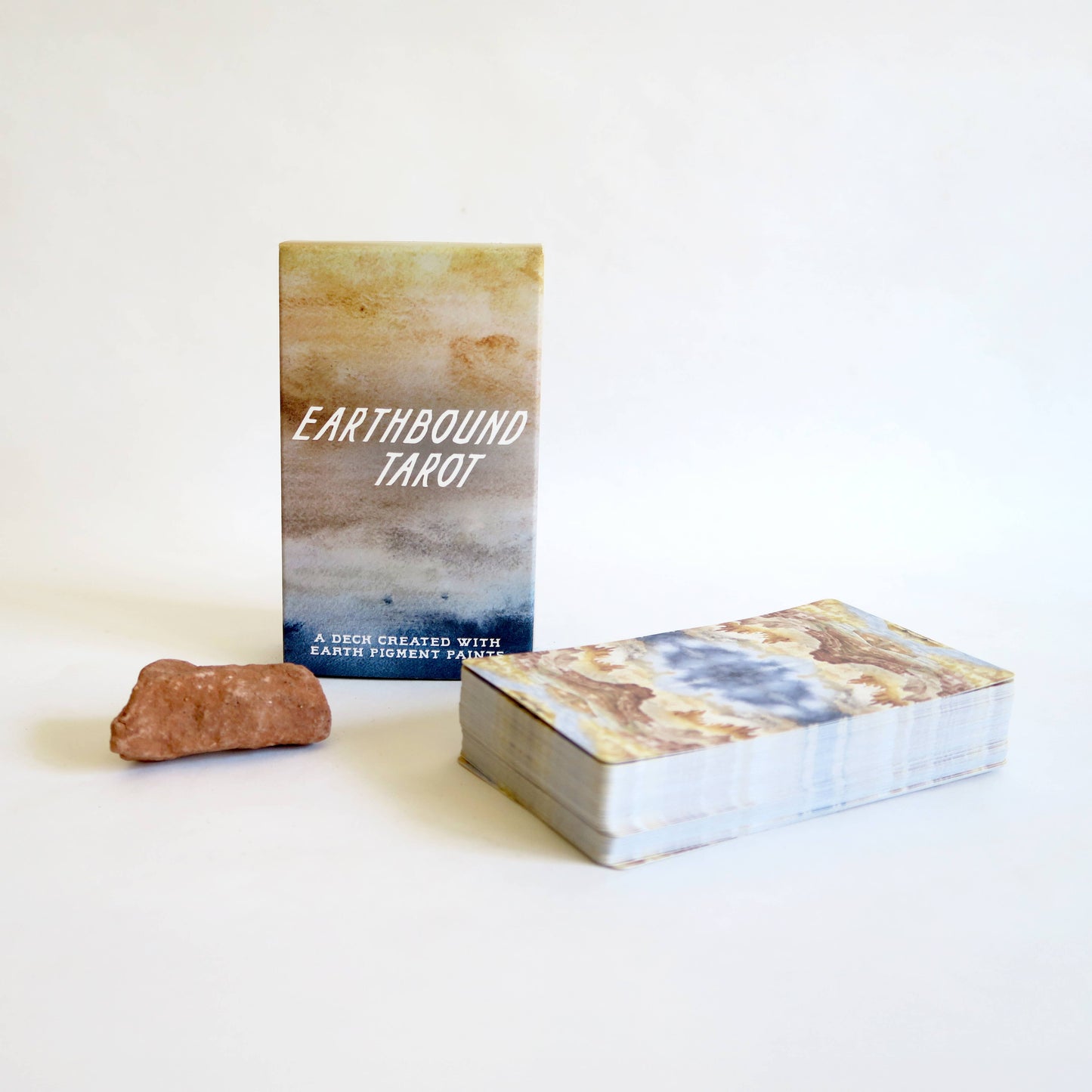 Earthbound Tarot deck standing upright next to a small brown crystal on a clean white background, with the deck of cards lying flat in front. The card back features an abstract marbled design in beige, brown, blue, and white.