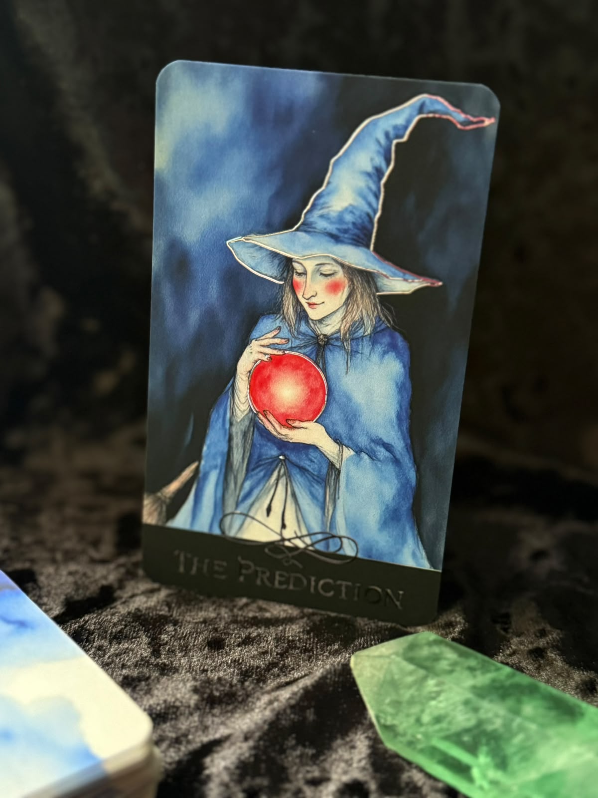 A tarot card titled 'THE PREDICTION' featuring an illustration of a blue-robed witch holding a glowing red orb, photographed standing upright on a dark textured surface with crystals nearby.