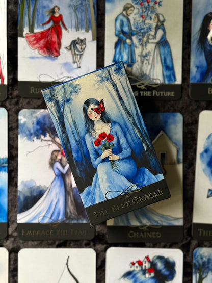 A tarot card deck box labeled 'THE BLUE ORACLE' is placed on top of several illustrated oracle cards featuring blue-toned women in dresses, a wolf, and floral scenes. The main product appears to be the oracle card deck itself rather than a lifestyle scene, so subject_type is Product/Packaging.