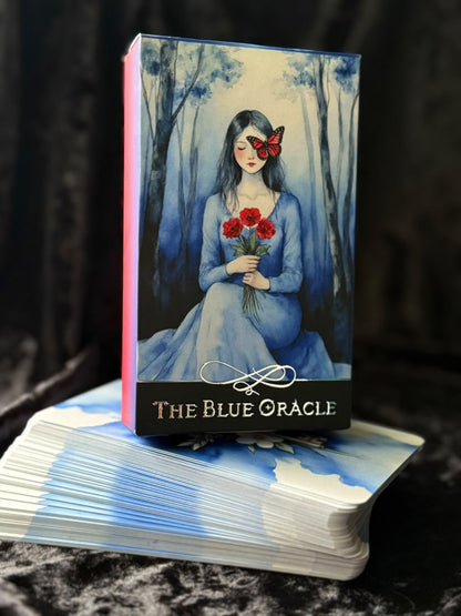 The Blue Oracle tarot deck box standing upright on top of a fanned deck of cards, with the front cover showing an illustrated woman in a blue dress holding red roses in a misty blue forest. The box has a pink spine and the title 'THE BLUE ORACLE' at the bottom.