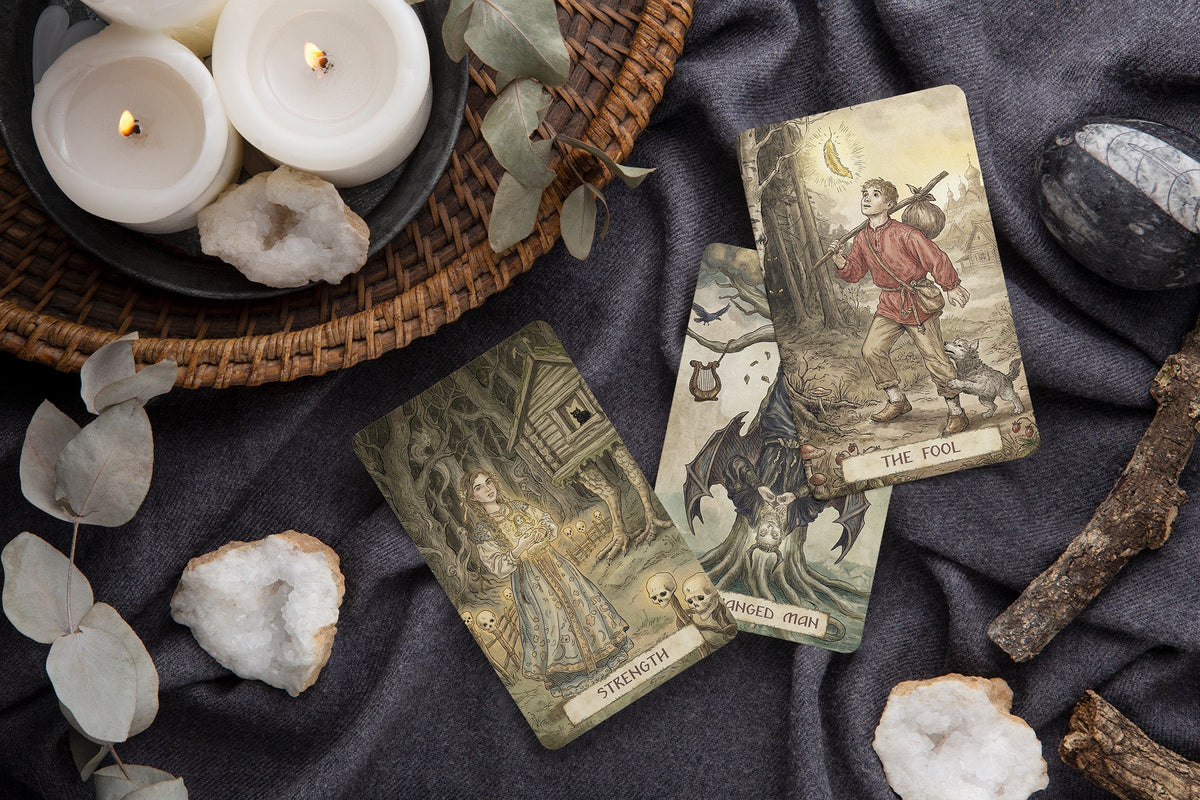 Three illustrated tarot cards are arranged on dark draped fabric with candles, crystals, eucalyptus leaves, and small branches around them. The visible card titles include The Fool, Strength, and Regained Power. Firebird Tarot | Slavic Folklore Indie Deck UK