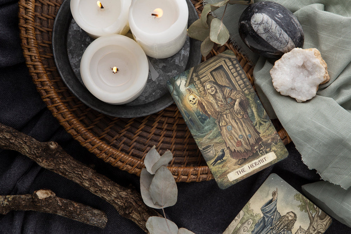 A tarot card deck styled in a moody, mystical flat-lay arrangement. The main card, 'The Lament', is displayed on a dark woven tray alongside three lit white pillar candles, a black stone, a white crystal, a sage green cloth, and dried eucalyptus leaves. Firebird Tarot | Slavic Folklore Indie Deck UK