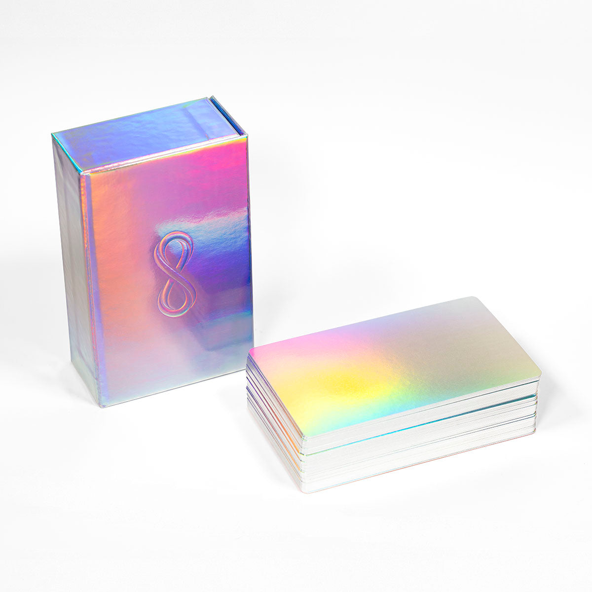 An iridescent holographic playing card deck with a matching box, featuring a metallic rainbow sheen and a raised infinity symbol on the box, set against a plain white background. Tarot of the Holy Spectrum Infinity Edition Holographic Mirror by Chase Voorhees, featuring colourful tarot cards, reflective holographic backs and edges, and a mirrored magnetic box.
