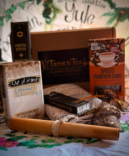 A curated gift box or subscription bundle containing various tea, scented candle, and book items arranged on a floral surface. Visible items include a box labeled 'Tarot & Tea', a candle labeled 'Crystal Scents', a book titled 'Out of Hand: Revisited', a spiced pumpkin chai tea box, and a rolled wooden or cork scroll tied with string.