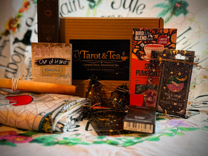 A curated tea and card gift set arranged on a patterned fabric surface, featuring a brown cardboard box, a Tarot & Tea box, a Bird Blend tea box, a Spice Pumpkin tea box, a rolled wooden paddle, wrapped cards, and small decorative items.