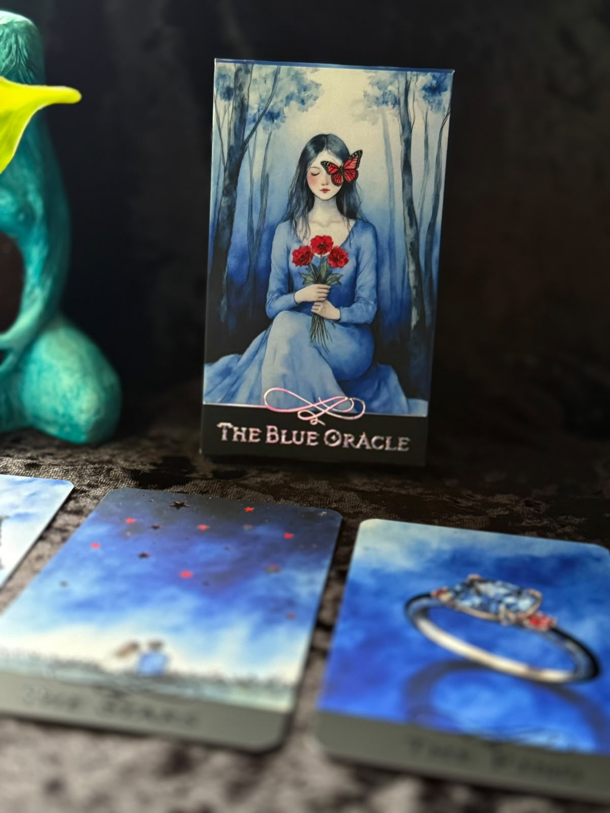 The Blue Oracle tarot deck box standing upright on a dark surface, with several illustrated oracle cards spread in front. The box art shows a seated woman in a blue dress holding red roses in a forest.
