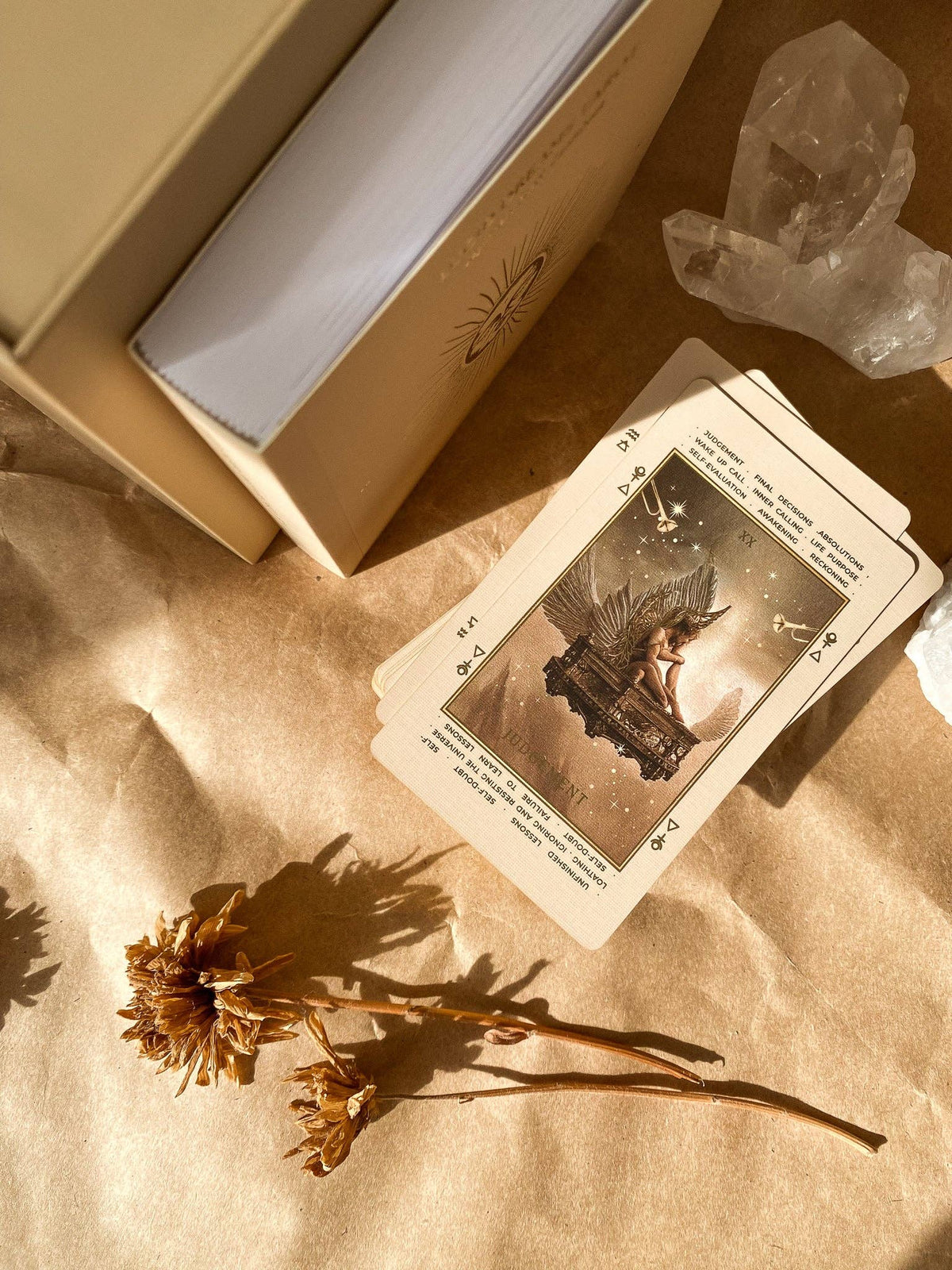 A tarot card deck with a beige box, shown with several cards laid out on parchment paper alongside dried flowers and a clear crystal cluster in warm sunlight. Lucid Dreams Beginner's Tarot Beige Edition IV by ST. SOLEIL with gold foil cards, matte gold edges and guidebook