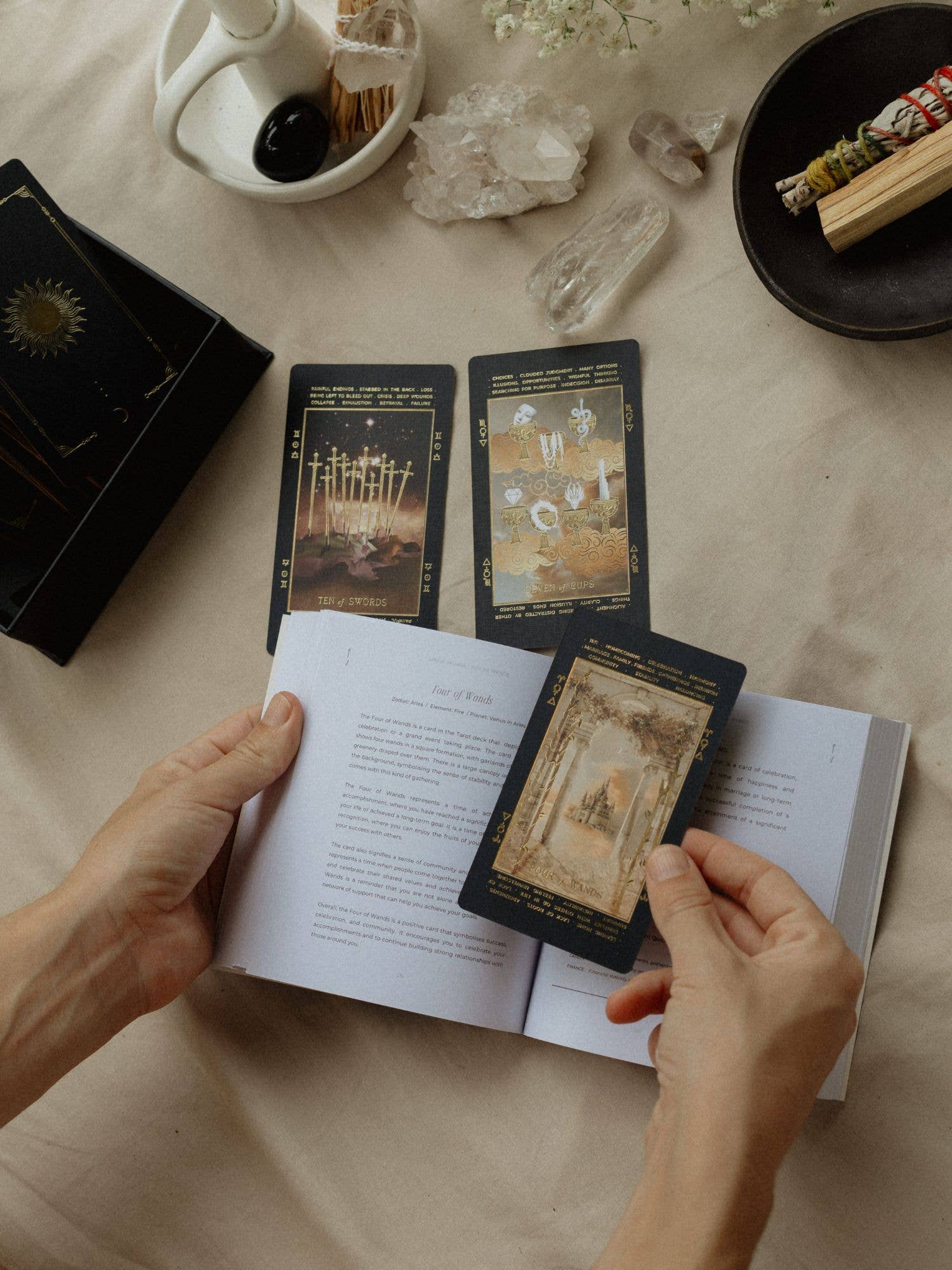 A person's hands holding an open tarot card book over a beige fabric surface, with several tarot cards spread out above it. The scene includes crystals, a white ceramic dish, and a black bowl with a smudge stick in the background.
