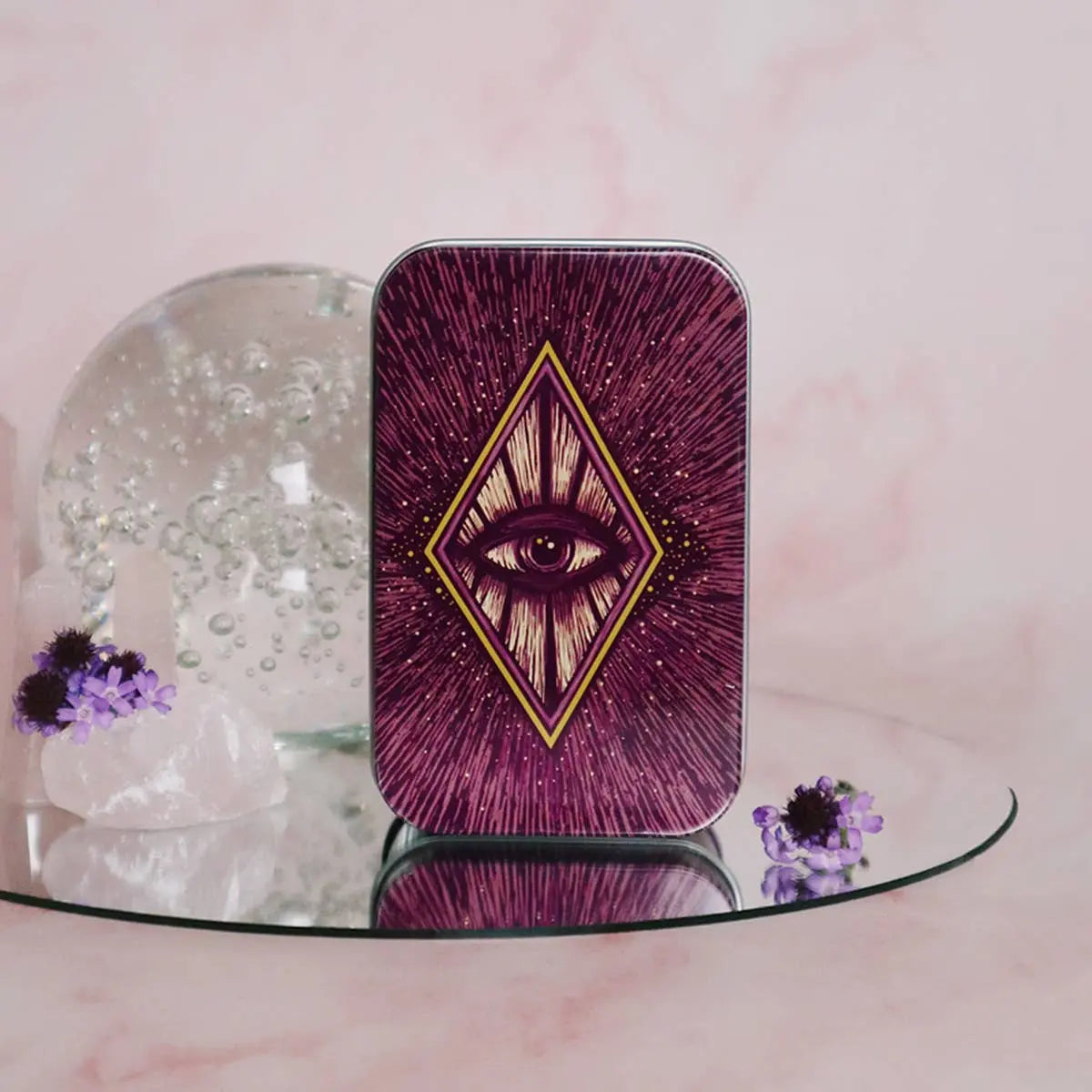Decorative tin with eye design on a pink surface with flowers and a crystal ball.