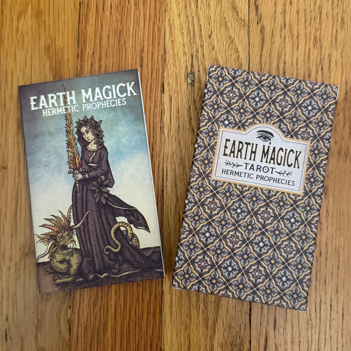 Two boxed tarot card decks from Earth Magick Hermetic Prophecies placed on a wooden tabletop. One box shows illustrated cover art with a robed figure and animal, and the other shows a patterned box front with the title label.