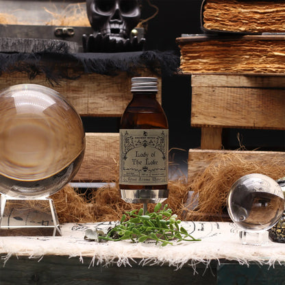 Bottle of 'Lady of the Lake' ritual water with a skull, books, and glasses on a wooden table.