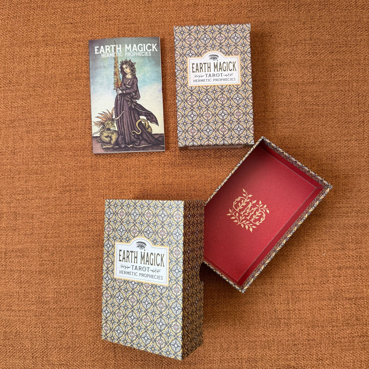 Earth Magick Tarot Card deck packaging shown on a brown textured fabric background, including the illustrated card, two patterned boxes, and an open box revealing a red interior with gold foil detailing.
