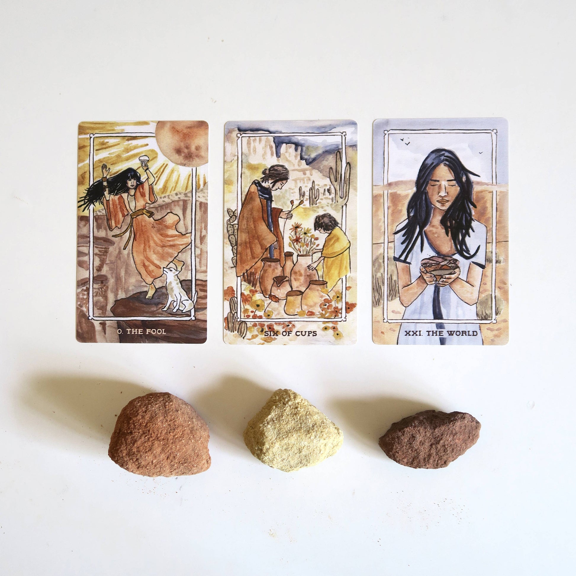 Three tarot cards with artistic illustrations of people and nature, accompanied by three stones on a white background.